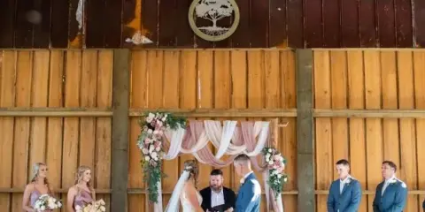 A bride and groom exchanging vows at a floral arch inside a wooden barn venue, flanked by bridesmaids and groomsmen.