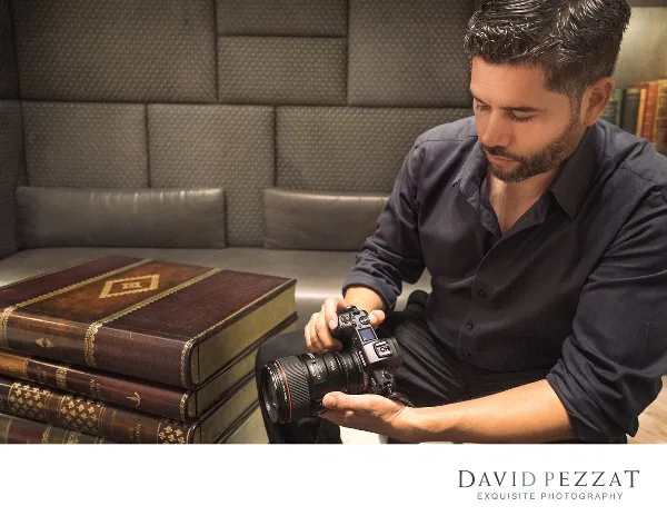 A professional photographer in a dark shirt holding a DSLR camera, seated next to a stack of leather-bound books.