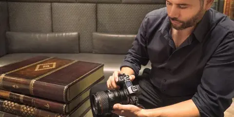 A professional photographer in a dark shirt holding a DSLR camera, seated next to a stack of leather-bound books.