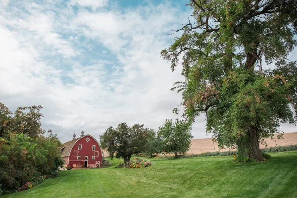 A red historic barn with white trim sits on a manicured green lawn with trees and flower beds under a cloudy sky.