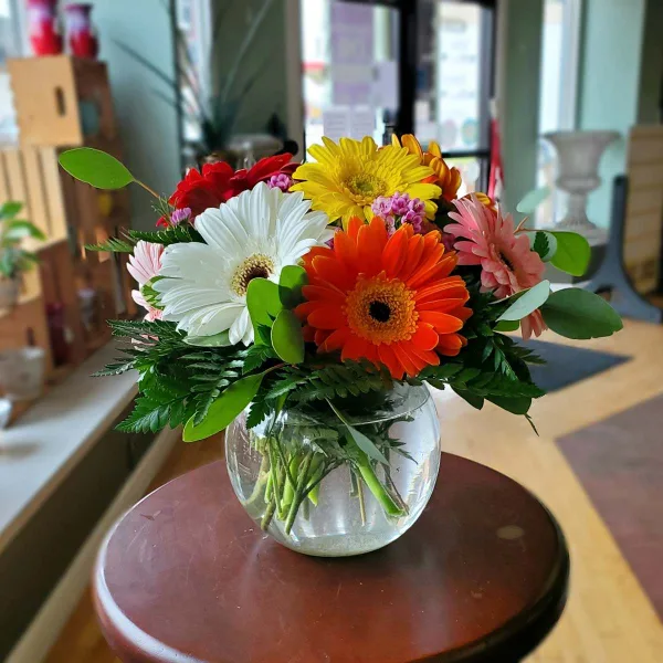 A colorful bouquet of gerbera daisies and greenery in a round glass vase on a wooden table.