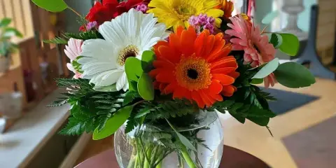 A colorful bouquet of gerbera daisies and greenery in a round glass vase on a wooden table.