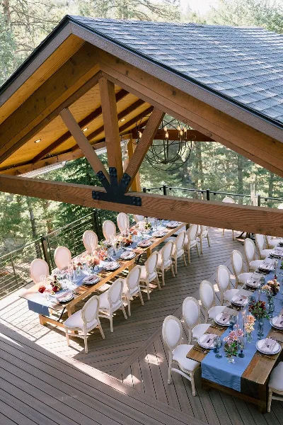 A rustic wooden wedding pavilion featuring long banquet tables set with floral arrangements and white chairs on a deck overlooking a forest.