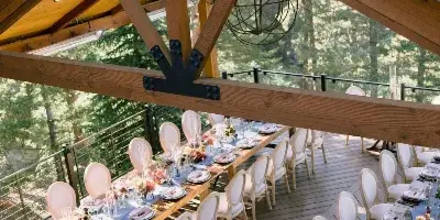 A rustic wooden wedding pavilion featuring long banquet tables set with floral arrangements and white chairs on a deck overlooking a forest.