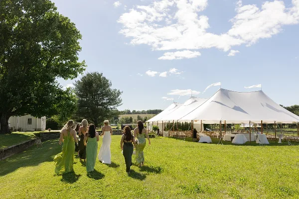 A wedding party walks across a green lawn toward a large white event tent set up with tables and chairs.
