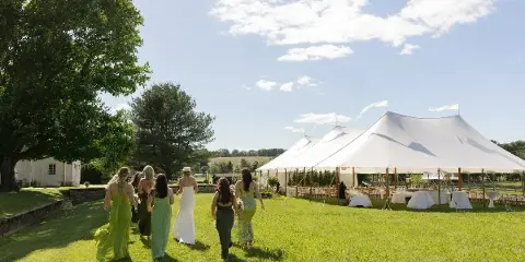 A wedding party walks across a green lawn toward a large white event tent set up with tables and chairs.