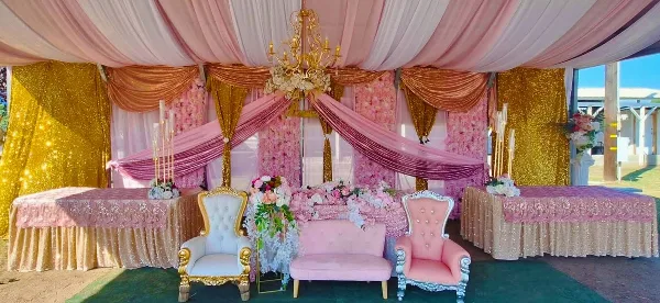 Decorated event space with pink and gold floral backdrops, sequin draping, chandeliers, and ornate chairs and tables.