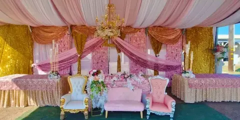 Decorated event space with pink and gold floral backdrops, sequin draping, chandeliers, and ornate chairs and tables.