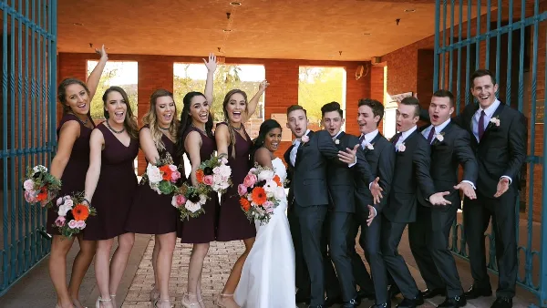 A wedding party consisting of bridesmaids in maroon dresses and groomsmen in dark suits posing playfully with a bride in a white dress.