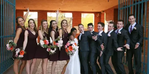 A wedding party consisting of bridesmaids in maroon dresses and groomsmen in dark suits posing playfully with a bride in a white dress.