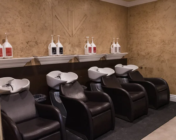 Interior of a hair salon showing a row of dark leather shampoo chairs with white basins, a textured wall with an NV logo, and shelves holding SEVEN hair care bottles.