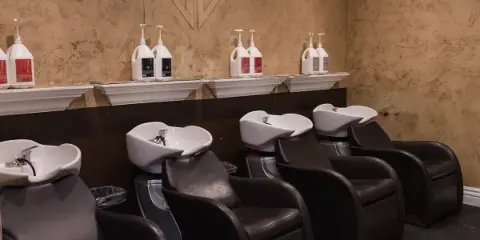Interior of a hair salon showing a row of dark leather shampoo chairs with white basins, a textured wall with an NV logo, and shelves holding SEVEN hair care bottles.