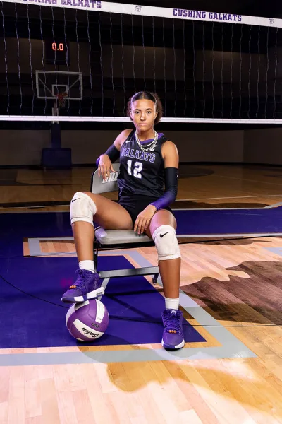 A female volleyball player in a black and purple uniform sits on a chair in a gymnasium with a volleyball at her feet.