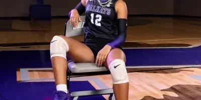 A female volleyball player in a black and purple uniform sits on a chair in a gymnasium with a volleyball at her feet.