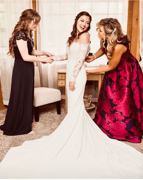 A bride in a white lace gown is being assisted by two women in a bright room with large windows.