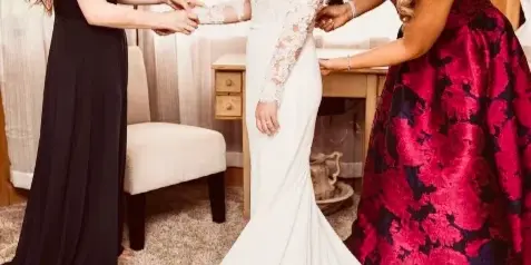 A bride in a white lace gown is being assisted by two women in a bright room with large windows.