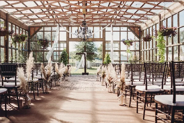 Interior view of a glass-walled greenhouse wedding venue with wooden beams, chandeliers, and rows of chairs set for a ceremony.