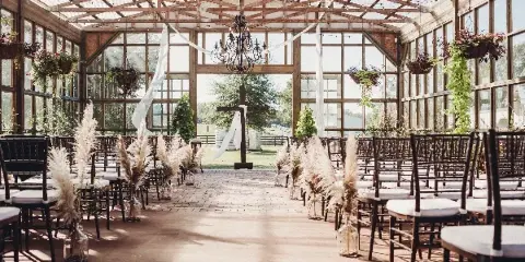 Interior view of a glass-walled greenhouse wedding venue with wooden beams, chandeliers, and rows of chairs set for a ceremony.