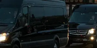 A line of black Mercedes-Benz passenger vans parked in a row with headlights on.