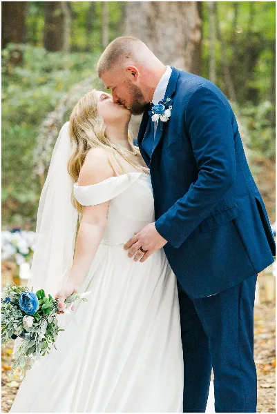 A bride in a white off-the-shoulder gown and a groom in a blue suit share a kiss in a wooded area.