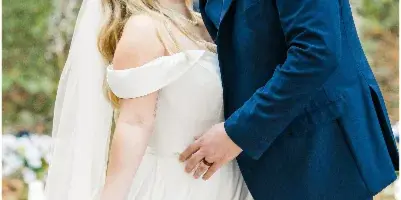 A bride in a white off-the-shoulder gown and a groom in a blue suit share a kiss in a wooded area.