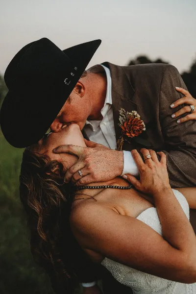 A bride and groom sharing a kiss, with the groom wearing a black cowboy hat and the bride in a white wedding dress.