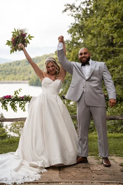 A bride in a white gown and a groom in a grey suit standing outdoors with arms raised, holding a bouquet, with a lake and trees in the background.
