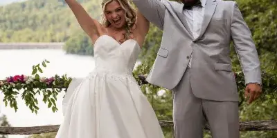 A bride in a white gown and a groom in a grey suit standing outdoors with arms raised, holding a bouquet, with a lake and trees in the background.