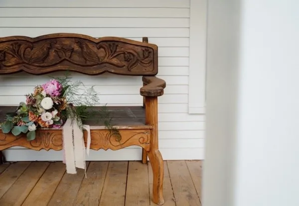 A carved wooden bench with a floral bouquet sits on a wooden porch against white siding.