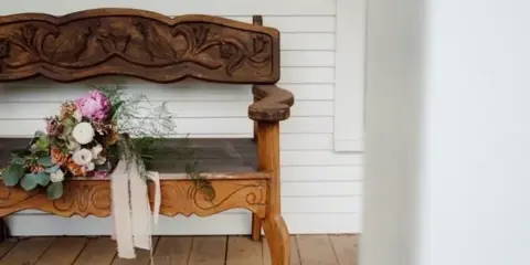 A carved wooden bench with a floral bouquet sits on a wooden porch against white siding.