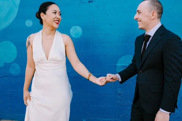 A bride in a white dress and a groom in a dark suit holding hands and smiling at each other against a vibrant blue wall.