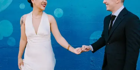 A bride in a white dress and a groom in a dark suit holding hands and smiling at each other against a vibrant blue wall.