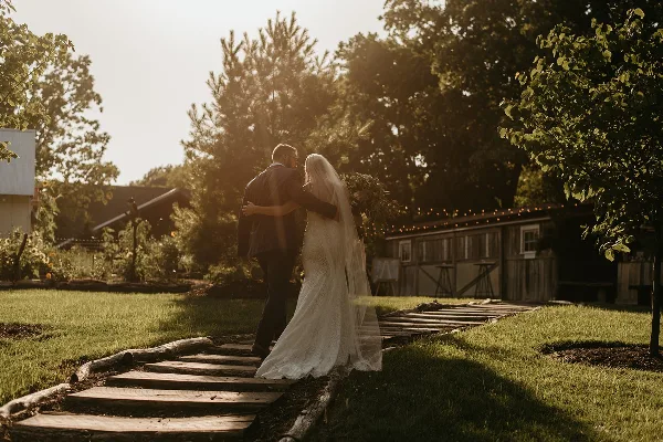 A bride and groom embracing on a wooden path in a grassy outdoor area with trees and a rustic building in the background.