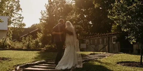A bride and groom embracing on a wooden path in a grassy outdoor area with trees and a rustic building in the background.