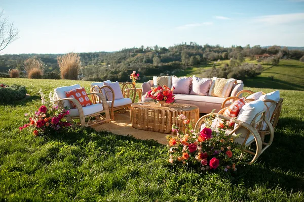 Outdoor wedding lounge set with wicker furniture, pink sofa, colorful floral arrangements, and a scenic vineyard landscape.