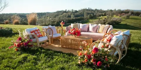 Outdoor wedding lounge set with wicker furniture, pink sofa, colorful floral arrangements, and a scenic vineyard landscape.