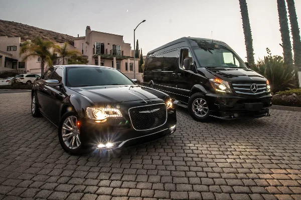 A black luxury sedan and a black Mercedes Sprinter van parked on a cobblestone driveway in front of a building.