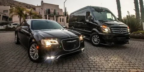 A black luxury sedan and a black Mercedes Sprinter van parked on a cobblestone driveway in front of a building.