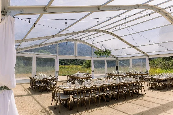 Interior of a clear wedding tent featuring long dining tables, wooden chairs, and string lights with a view of a grassy field and hills.
