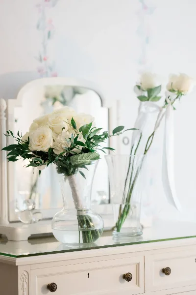 A vase of white roses and greenery sitting on a white dresser with a mirror in the background.