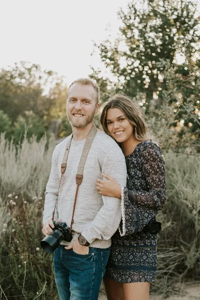 A smiling man and woman standing outdoors, both holding professional DSLR cameras.
