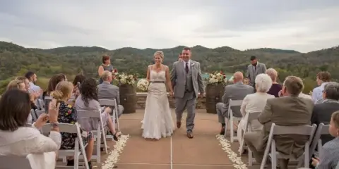 A bride and groom walking down the aisle during an outdoor wedding ceremony with guests seated on either side.