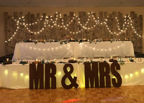 A wedding reception venue featuring a head table with a 'Mr & Mrs' sign, white table linens, and string lights against a wooden backdrop.
