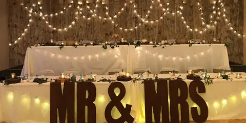 A wedding reception venue featuring a head table with a 'Mr & Mrs' sign, white table linens, and string lights against a wooden backdrop.