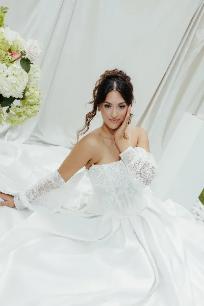 A bride wearing a white lace gown with a styled updo and professional makeup, posing next to a floral arrangement.
