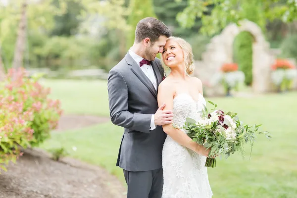 A bride and groom standing together in a garden, with the groom embracing the bride who is holding a bouquet.