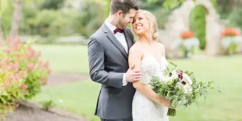 A bride and groom standing together in a garden, with the groom embracing the bride who is holding a bouquet.