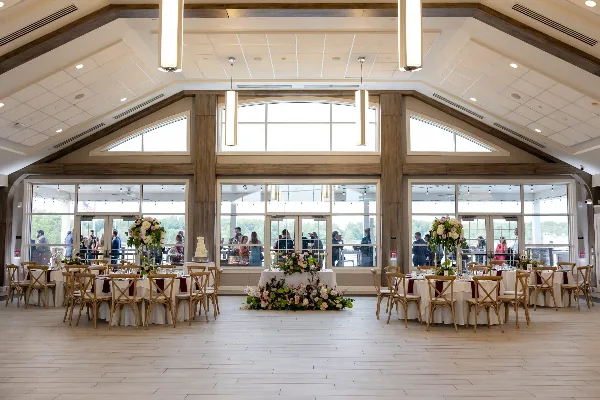A spacious indoor event venue with round tables set for a wedding reception, featuring floral arrangements and a cake.