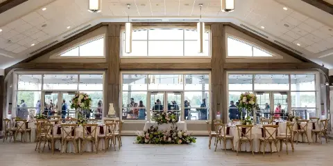 A spacious indoor event venue with round tables set for a wedding reception, featuring floral arrangements and a cake.