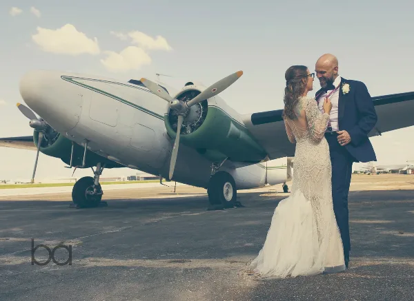 A bride and groom standing together in front of a vintage propeller airplane.